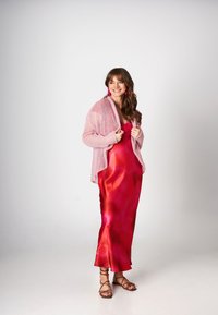Kobieta w błyszczącej czerwonej satynowej sukience o wirujących tonach, nałożonej na miękki różowy kardigan. Ma na sobie różowe bijące w oczy kolczyki i sandały na cienkich paskach.