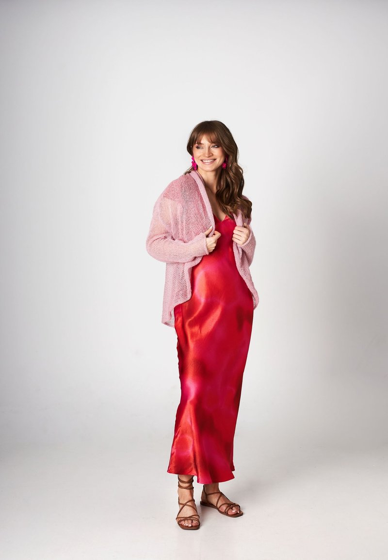 Kobieta w błyszczącej czerwonej satynowej sukience o wirujących tonach, nałożonej na miękki różowy kardigan. Ma na sobie różowe bijące w oczy kolczyki i sandały na cienkich paskach.