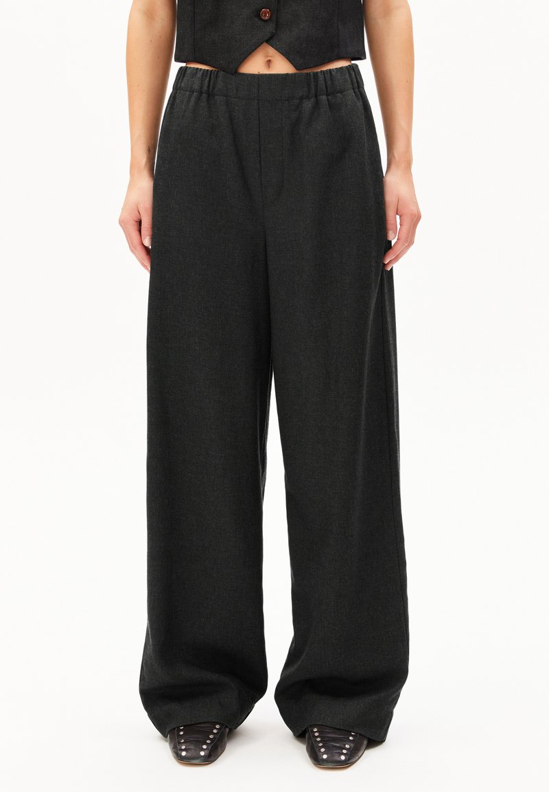 Breda byxor i mörkgrått tyg, med ett elastiskt midjeband för komfort. Slät textur utan synliga mönster; i kombination med spetsiga skor.