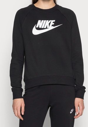 Person iført sort Nike-sweatshirt og matchende sorte bukser, begge med hvide Nike-logoer, stående mod en ensfarvet baggrund.