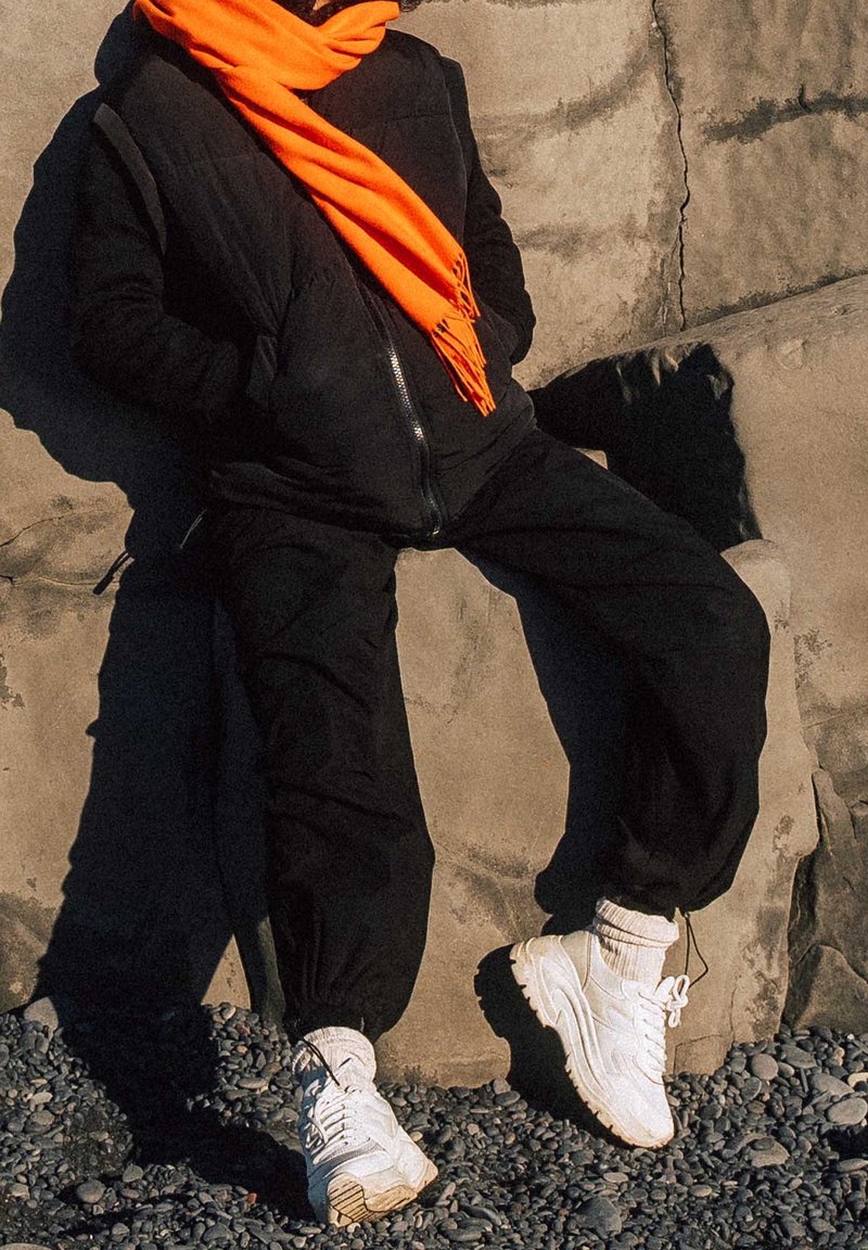 Personne appuyée contre un mur en pierre portant un pantalon noir, une veste noire, une écharpe orange vif, des chaussettes blanches et des baskets blanches épaisses sur un sol en gravier.