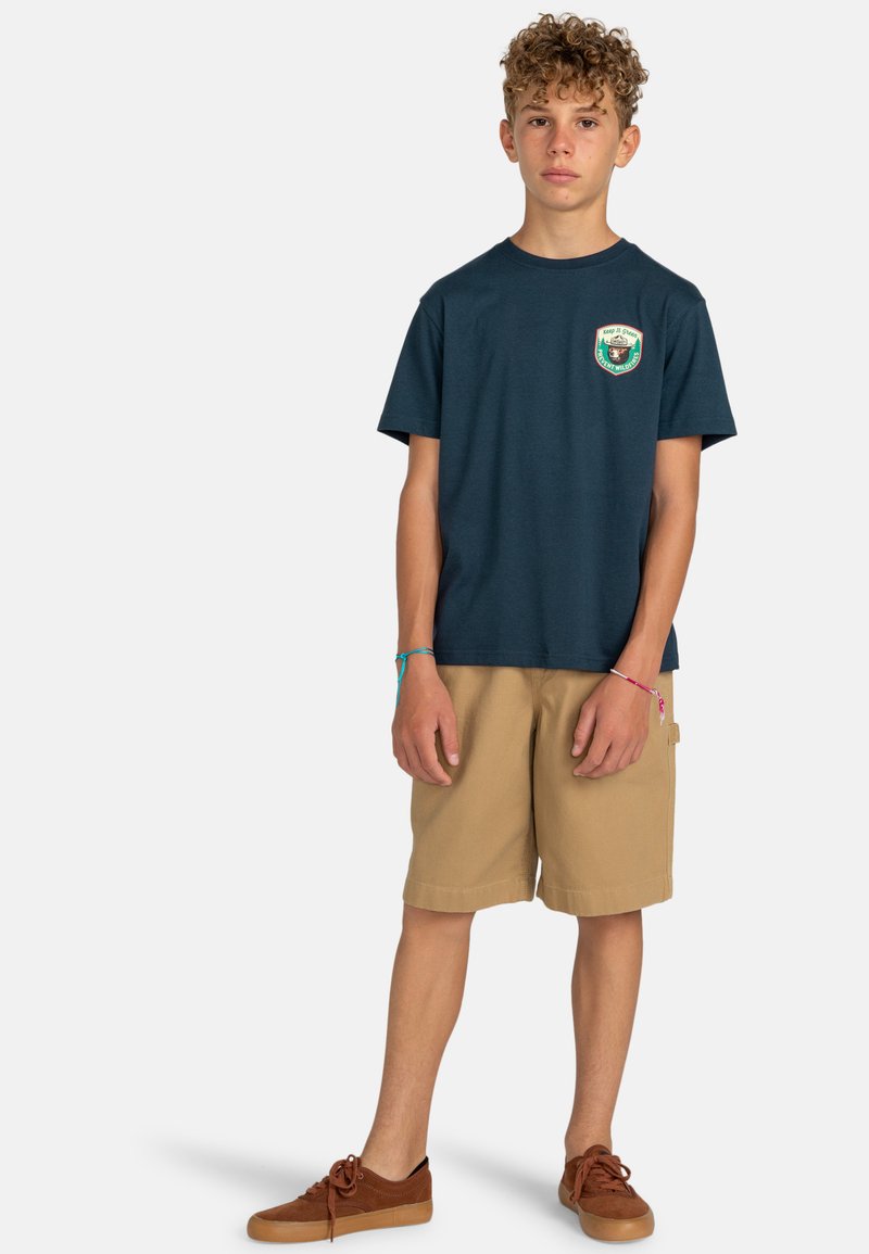 T-shirt en coton bleu marine à manches courtes, ornée d'une écusson logo coloré sur la poitrine, associé à un short beige et des chaussures marron.