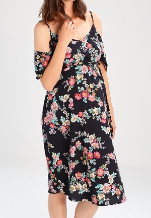 Kobieta w czarnej, sięgającej do kolan sukience w kwiaty, z odkrytymi ramionami i cienkimi ramiączkami, stoi na jednolitym tle.