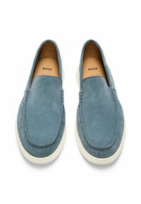 Scarpe slip-on in suede blu con punta tonda, suola in gomma bianca e dettagli di cucitura minimi. Il marchio è visibile sulle solette.