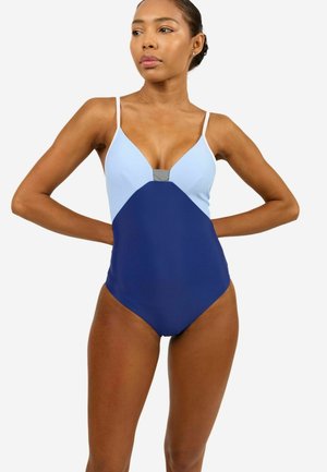 Woman wearing a sleeveless, color-block one-piece swimsuit with light blue top and navy blue bottom, standing with hands behind back.