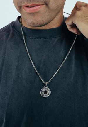 Collier en argent avec un pendentif circulaire présentant un design en éclat de soleil texturé et une pierre noire centrale. Porté sur une chemise sombre.