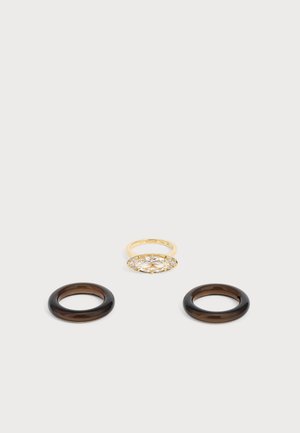 Anillo de oro con una piedra preciosa ovalada y clara rodeada de pequeñas piedras, mostrado sobre dos bandas circulares marrón oscuro sobre un fondo claro.