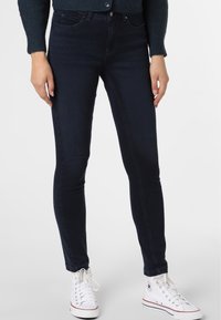 Persona con jeans skinny oscuros, zapatillas blancas de caña alta y un suéter azul oscuro, de pie con las manos relajadas a los lados.