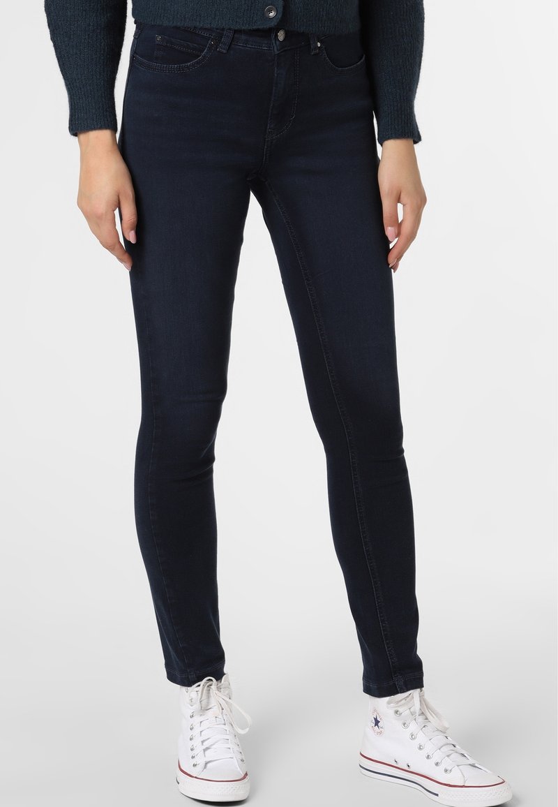 Persona con jeans skinny oscuros, zapatillas blancas de caña alta y un suéter azul oscuro, de pie con las manos relajadas a los lados.