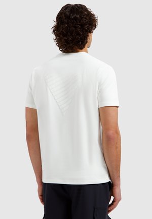 Man met krullend haar die een eenvoudige witte T-shirt met korte mouwen draagt met een subtiel driehoekig patroon op de bovenrug, en zwarte broek.