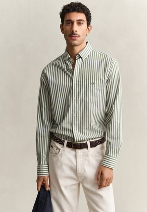 Man wearing green and white vertical striped button-up shirt, beige pants, brown belt, holding dark blue jacket against plain background.