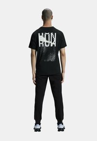 T-shirt noir avec un grand texte graphique blanc au dos. Associé à un pantalon noir et des chaussures de sport grises. À manches courtes, coupe décontractée.