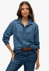 Vrouw in lange mouwen blauwe denim shirt met twee borstzakken en een gevlochten bruine riem, gecombineerd met blauwe jeans, poserend tegen een witte achtergrond.