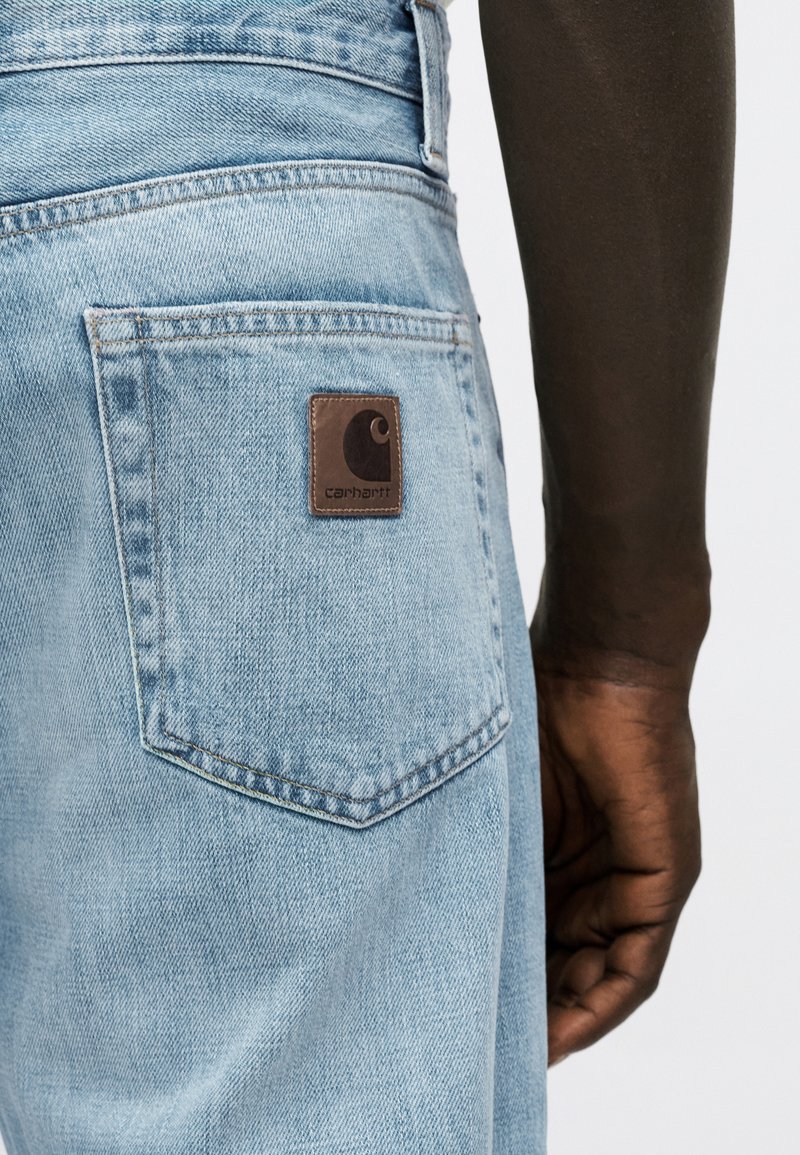 Light blue denim jeans pocket with brown Carhartt logo patch, worn by a person with dark skin and a relaxed hand positioned beside the jeans.