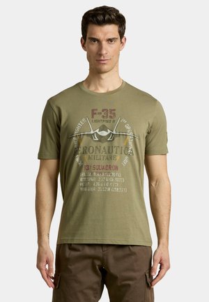 Uomo che indossa una t-shirt verde oliva con grafica del jet F-35 e testo sull'Aeronautica Militare e il 13º Stormo, in piedi davanti a uno sfondo semplice.