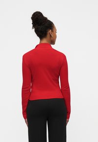 Frau mit lockigem Haar im Dutt, trägt ein rotes Langarmshirt und schwarze Hose, von hinten vor einem schlichten Hintergrund gezeigt.