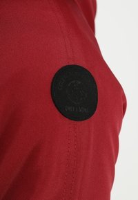Tela roja con una textura suave que presenta un parche circular negro con la inscripción "ONLY & SONS." El parche tiene textura, añadiendo contraste visual.