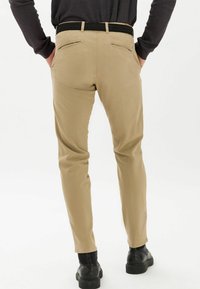 Beżowe spodnie o wąskim kroju, z dwoma tylnymi kieszeniami i czarnym plecionym paskiem, zestawione z czarnymi butami, widoczne od tyłu.