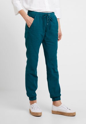 Pantalon de jogging bleu sarcelle à taille élastique avec cordon et poches latérales, porté avec des baskets blanches et un haut blanc à manches longues.