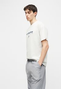 Νεαρός άνδρας με κοντά σκούρα μαλλιά φορώντας λευκό μπλουζάκι "New Balance Athletics" και ανοιχτό γκρι παντελόνι με τα χέρια στις τσέπες, κοιτάζοντας αριστερά.