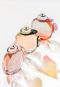 Tre parfymeflasker: rosa, transparent og oransje. Hver har en rund form, gullkapsler og distinkte pregete design med fargerike aksenter.