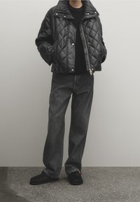 Osoba ubrana w czarną pikowaną kurtkę na czarnej koszuli, luźne ciemne dżinsy i czarne buty wsuwane stojąca na tle jednolitego tła.