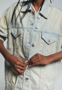 Chaqueta de denim azul claro con un corte relajado, que presenta botones plateados, dos bolsillos en el pecho y detalles deshilachados en la tela.