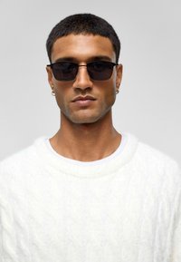 Schwarze rechteckige Sonnenbrille mit dünnem Rahmen, getragen von einer Person in einem weißen Strickpullover. Glatte Textur, minimalistisches Design und keine Verzierungen.