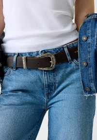 Ceinture en cuir noir avec une boucle argentée ornée, portée au-dessus d'un jean en denim bleu et d'un débardeur blanc côtelé. Une veste en denim est partiellement visible.