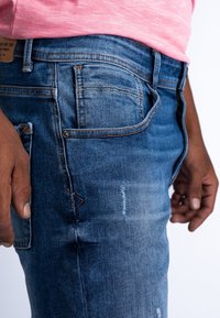 Vaqueros de mezclilla en azul medio con un acabado desgastado. Las características incluyen costuras cosidas, un pequeño bolsillo y detalles de desgaste en el muslo.