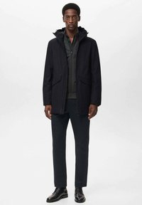 Chaqueta con capucha negra y dos bolsillos frontales, combinada con una camiseta negra, pantalones oscuros y zapatos negros. Textura suave, diseño minimalista.