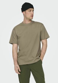 Camiseta de algodón color caqui con cuello redondo y mangas cortas, combinada con pantalones verde oliva. El modelo lleva un gorro negro acanalado.