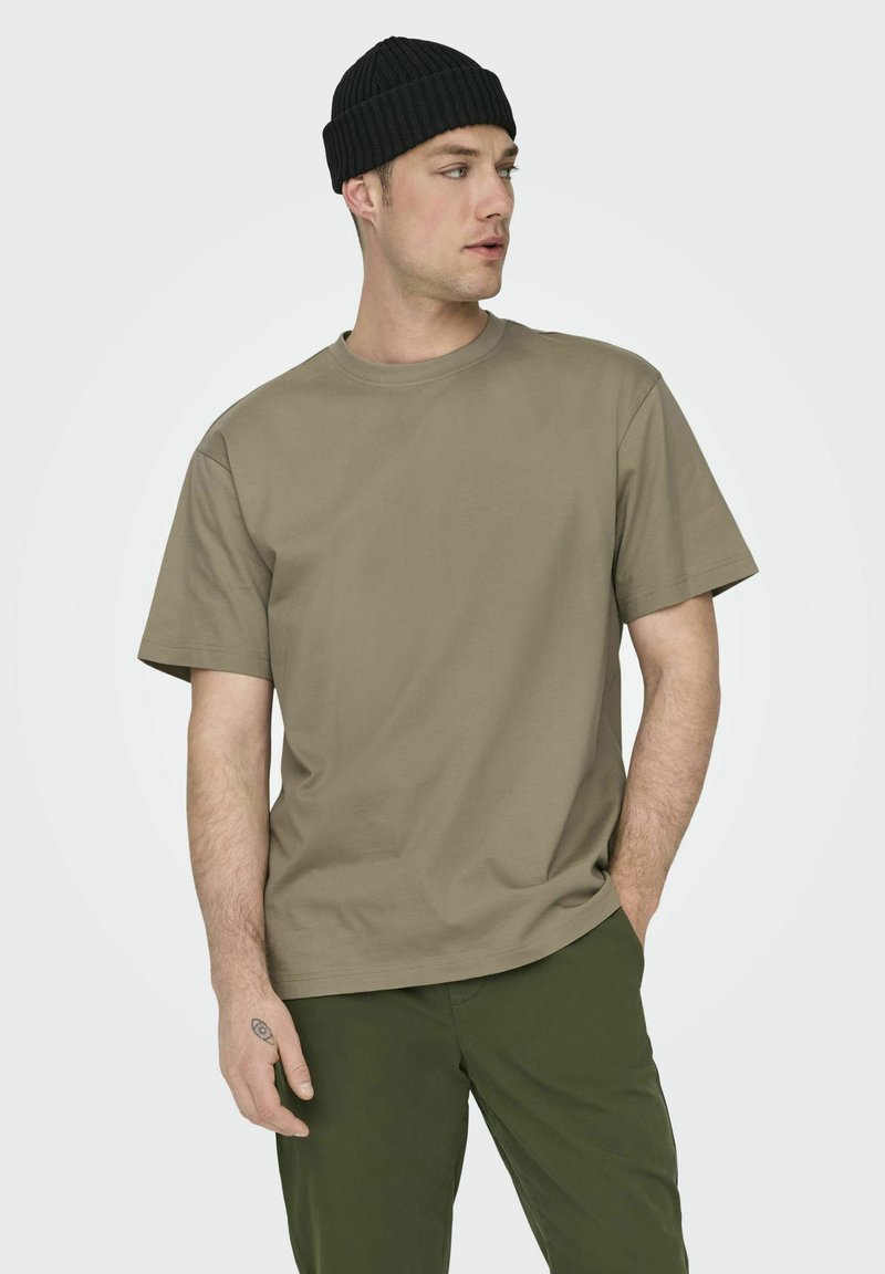 Khaki katoenen t-shirt met een ronde hals en korte mouwen, gecombineerd met olijfgroene broek. Het model draagt een zwarte geribde beanie.