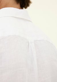 Camicia bianca in lino con una texture morbida, caratterizzata da un colletto classico e un design a yoke sul retro, che presenta un leggero dettaglio a pieghe.