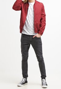 Giubbotto bomber rosso, camicia bianca con piccoli motivi neri, jeans slim-fit grigi, sneaker alte nere. Tessuto morbido, design casual.