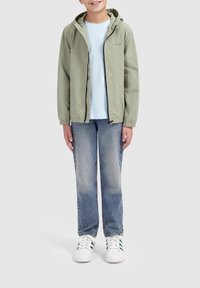 Lichtgroene, ritsjack met capuchon, elastische manchetten, gedragen over een lichtblauw shirt. In combinatie met blauwe spijkerbroeken en witte sneakers met groene accenten.