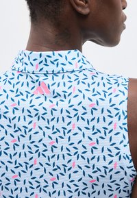 Άτομο που φοράει χωρίς μανίκια πουκάμισο με γιακά, με μπλε και ροζ μοτίβο φύλλων και ροζ λογότυπο Adidas στο πάνω μέρος της πλάτης.