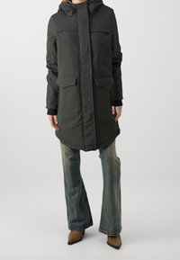 Lång mörkgrön parkas med huva, som har två framfickor, rak skärning och kontrasterande svarta detaljer på ärmsluten. Bärs över utsvängda jeans.