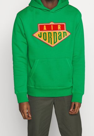 Mężczyzna w zielonej bluzie z kapturem z czerwonym, żółtym i zielonym logo Air Jordan, w ciemnych spodniach, stojący na jednolitym tle.