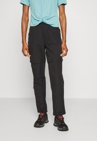 Svarta zip-off byxor med en strukturerad yta, med sidfickor och logotyp. I kombination med en ljusblå knuten t-shirt och svarta skor.