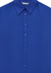 Chemise bleue à manches longues avec boutons, col pointu et étiquette de marque visible cousue à l'intérieur de l'encolure.