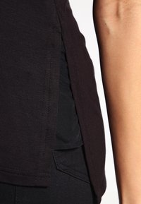 Camicia di cotone nera con fessure laterali, maniche corte e una trama liscia. È indossata sopra un indumento nero, evidenziando un design aderente.