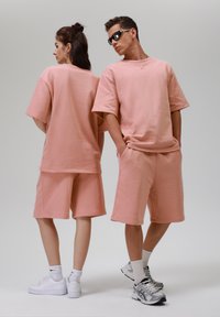 Růžové bavlněné oversized mikiny a šortky s uvolněným střihem. Oba modely stojí zády k sobě a mají na sobě bílé a šedé sportovní boty.
