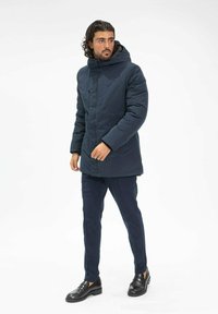Manteau rembourré navy avec capuche, présentant un zip à l'avant, une finition texturée et des manches longues. Associé à un pantalon navy et des chaussures noires.