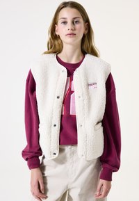 Elfenbeinfarbene, flauschige Weste mit Druckknöpfen über einem burgunderfarbenen Sweatshirt, das eine große Zahl und ein Logo zeigt. Lässiger Schnitt, mit einer Tasche auf jeder Seite.
