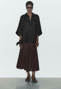 Schwarzes, übergroßes Hemd mit Knöpfen und lockeren Ärmeln, kombiniert mit einem fließenden, braunen Faltenrock und schwarzen Riemchensandalen. Stylisierte Sonnenbrille inklusive.