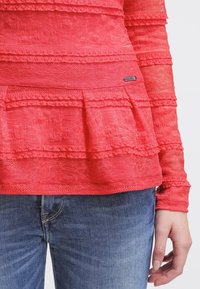 Top de manga larga color coral con detalles de encaje en capas y un dobladillo peplum. Etiqueta visible en la cintura; combinado con jeans de mezclilla azules.