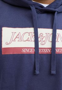 Marine hættetrøje med et cremefarvet og maroon logo grafik med teksten "JACK&JON" og "SINCE NINETEEN". Indeholder snore og et tekstureret stof.