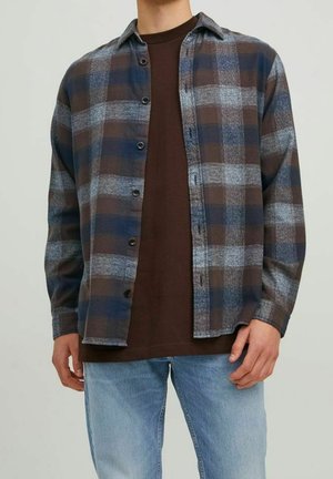 Mann trägt ein blaues, braunes und graues kariertes Hemd über einem dunkelbraunen T-Shirt mit hellblauen Jeans, steht vor einem schlichten Hintergrund.