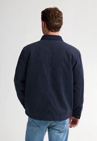 Middelblauwe jas met een kraag, knopenmanchetten en een ontspannen pasvorm. Zacht, gestructureerd materiaal met een strakke uitstraling en geen zichtbare patronen.